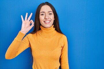 Sticker - Young brunette woman standing over blue background smiling positive doing ok sign with hand and fingers. successful expression.