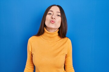 Sticker - Young brunette woman standing over blue background looking at the camera blowing a kiss on air being lovely and sexy. love expression.