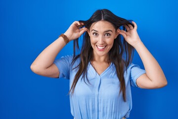 Poster - Young brunette woman standing over blue background smiling pulling ears with fingers, funny gesture. audition problem