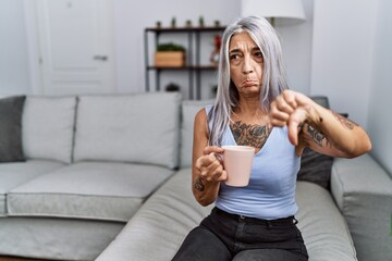 Sticker - Middle age grey-haired woman drinking coffee sitting on the sofa at home pointing down looking sad and upset, indicating direction with fingers, unhappy and depressed.