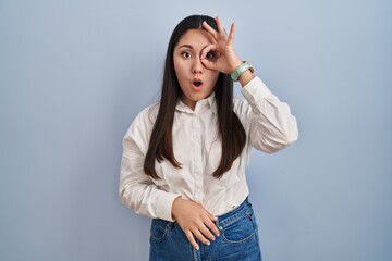 Sticker - Young latin woman standing over blue background doing ok gesture shocked with surprised face, eye looking through fingers. unbelieving expression.