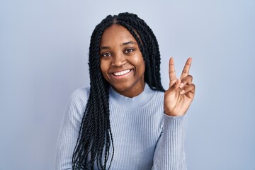 Wall Mural - African american woman standing over blue background smiling with happy face winking at the camera doing victory sign with fingers. number two.