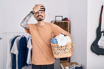 Wall Mural - Young hispanic man with beard holding laundry basket at bedroom stressed and frustrated with hand on head, surprised and angry face