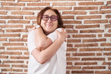 Poster - Senior woman with glasses standing over bricks wall hugging oneself happy and positive, smiling confident. self love and self care