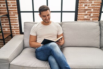 Wall Mural - Young hispanic man reading book sitting on sofa at home