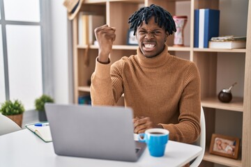 Poster - Young african man with dreadlocks working using computer laptop angry and mad raising fist frustrated and furious while shouting with anger. rage and aggressive concept.