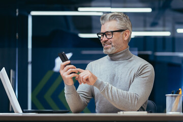 Mature successful businessman working inside office, senior gray-haired businessman using phone contentedly reading online message, man working with laptop at workplace, happy investor.