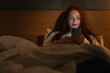 © dikushin - Portrait of happy smiling young woman using smartphone, looking on screen, typing online message on social media, lying on bed late at night, bedside lamp lighting with warm yellow light.