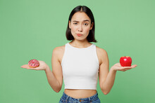 Young Pensive Sad Woman Wear White Clothes Hold In Hand Red Apple Pink Donut Look Camera Isolated On Plain Pastel Light Green Background. Proper Nutrition Healthy Fast Food Unhealthy Choice Concept.
