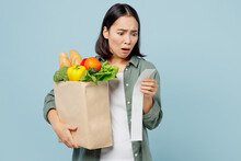 Young Confused Sad Woman Wearing Casual Clothes Hold Brown Paper Bag With Food Products Look At Check Isolated On Plain Blue Cyan Background Studio Portrait. Delivery Service From Shop Or Restaurant.