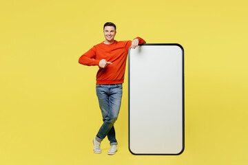 Wall Mural - Full body young man in orange casual clothes stand near big huge blank screen mobile cell phone point index finger on smartphone with copy space mockup area isolated on plain yellow color background