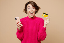 Young Woman Wear Pink Sweater Using Mobile Cell Phone Hold Credit Bank Card Doing Online Shopping Order Delivery Booking Tour Isolated On Plain Pastel Light Beige Background. People Lifestyle Concept.