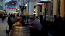 Defocused View Of Cozy Outdoor Restaurant At Evening. Unidentified People Eating Food And Having Conversation In An Outdoor Restaurant. Blurred Real Time Scene. People Having Fun And Drinking Wine.