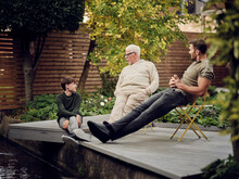 Father, Son And Grandson Sitting At Garden Pond Talking And Relaxing