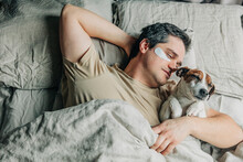 Man With Under Eye Patches Sleeping With Dog On Bed
