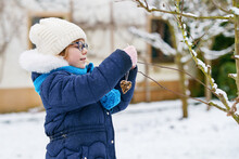 Little Girl With Glasses Feeds Birds On Winter. Happy Smiling Preschool Child Hanging Selfmade Bird Seed Heart On Tree. On Cold Snowy Winter Day.