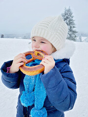 Wall Mural - Little preschool girl eating pretzel in winter forest. Happy healthy child in warm winter clothes, outdoors. Active family leisure, walking in snow landscape.