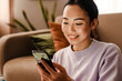© Drobot Dean - Cheerful asian woman using smart phone while sitting in living room