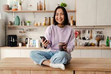 Wall Mural - Young smiling asian woman using mobile phone and drinking coffee in cozy kitchen