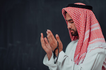 Arabian man in traditional clothes making traditional prayer to God, keeps hands in praying gesture in front of black chalkboard representing modern islam fashion and ramadan kareem concept