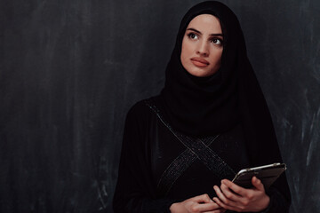 Wall Mural - Young modern muslim business woman using tablet computer wearing glasses and hijab clothes in front of black chalkboard