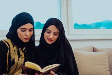 Wall Mural - Two Young traditional Muslim women read Quran on the sofa before iftar dinner during a Ramadan feast at home