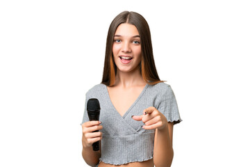 Sticker - Teenager singer girl picking up a microphone over isolated background surprised and pointing front