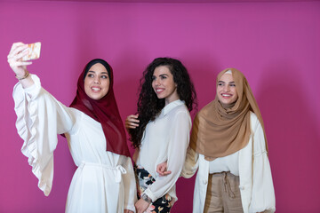 Wall Mural - Group of beautiful muslim women two of them in fashionable dress with hijab using mobile phone while taking selfie picture isolated on pink background representing modern islam fashion technology 