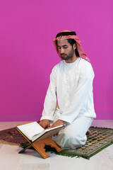 Wall Mural - Young arabian muslim man in traditional clothes reading holy book Quran on the praying carpet before iftar dinner during a ramadan feast at home