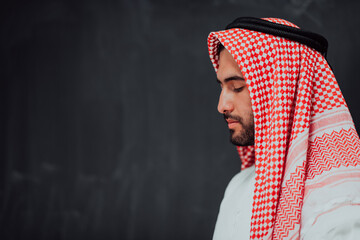 Wall Mural - Arabian man in traditional clothes making traditional prayer to God, keeps hands in praying gesture in front of black chalkboard representing modern islam fashion and ramadan kareem concept