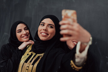 Wall Mural - Group of young beautiful muslim women in fashionable dress with hijab using smartphone while taking selfie picture in front of black background