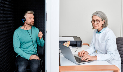 Wall Mural - Audiological exam. Audiologist doing hearing check-up to handsome adult man in soundproof audiometric booth. Hearing loss treatment, side view