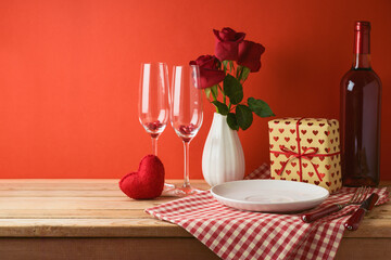 Wall Mural - Empty white plate with wine glasses, heart shape and rose flowers on wooden table over red background. Valentine's day romantic dinner concept