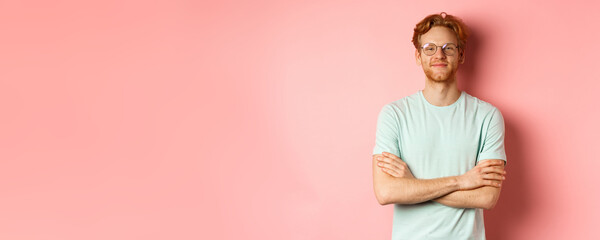 Portrait of satisfied caucasian man with red hair and beard, cross arms on chest and smiling with smug face, wearing glasses, standing over pink background