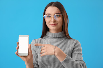 Poster - Young woman in eyeglasses pointing at mobile phone on blue background