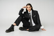 © LIGHTFIELD STUDIOS - full length of young model in black elegant pantsuit and rough boots sitting with hand near head on grey background.