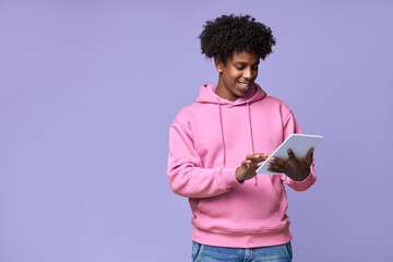 Young happy cool curly African American teenager student boy wearing pink hoodie holding pad using digital tablet computer technology browsing, elearning standing isolated on light purple background.