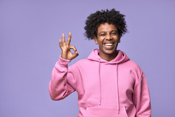 Wall Mural - Young happy hipster African American teen guy student wearing pink hoodie isolated on purple background. Smiling cool joyful ethnic generation z teenager model laughing showing ok sign and winking.