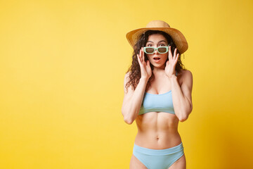 Wall Mural - young shocked brunette girl in a blue swimsuit and with curly hair holds sunglasses on a yellow background