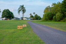Two Yellow Wooden Signs, No Carts And Cart Path Only This Hole, On Green Grass Next To A Paved Cart Path On A Golf Course. It's A Sunny Summer's Day With Palm Trees, Woods And Trailers Along The Edge.