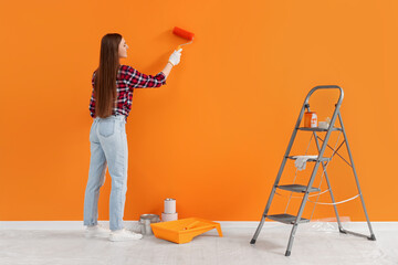 Wall Mural - Designer painting orange wall with roller near folding ladder indoors