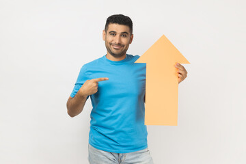 Portrait of cheerful unshaven man wearing blue T- shirt standing pointing at carton arrow indicating up, being glad of career success. Indoor studio shot isolated on gray background.