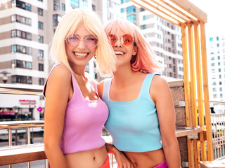 Two young beautiful smiling hipster female in trendy summer clothes. Sexy carefree women posing in the street in wigs. Positive funny models having fun at sunset. In sunglasses. At sunny day