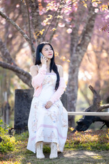 Wall Mural - Beautiful woman using smartphone, talking, typing message, chatting, scrolling web page on mobile phone under the pink blossoms cherry tree. Woman selfie and look at camera on sakura background.