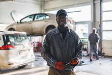Portrait Of An Auto Mechanic Holding A Metallic Wrench In A Busy Auto Repair Shop. High-quality Photo
