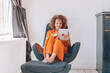 smiling, elegant lady with curly hair sitting barefoot on lounge chair in her home, scrolling through her tablet while basking in the natural light streaming through the nearby window