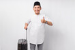 © Bangun Stock Photo - Cheerful young Asian Muslim man standing while holding suitcase in hand, showing thumb up gesture, ready for the holiday eid Mubarak isolated on white background. Ramadan and eid Mubarak concept