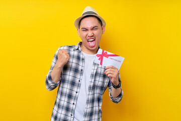 Wall Mural - Excited handsome Asian ethnicity young man wearing casual clothes hat holding gift certificate coupon voucher card and showing winner gesture isolated on yellow background. People lifestyle concept