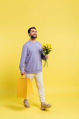 Wall Mural - full length of happy and stylish man walking with shopping bags and flowers on yellow background.
