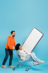 Wall Mural - bearded man laughing near shopping cart with african american woman and mobile phone template on blue background.